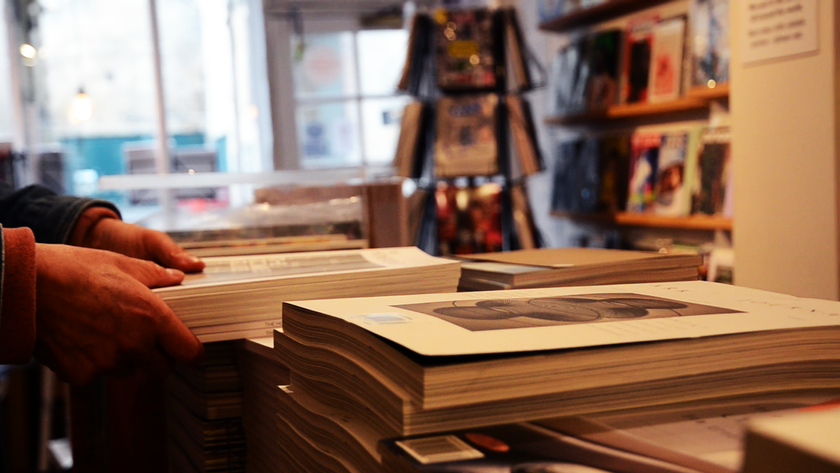 Photograph of a man moving a pile of independent magazines around a warmly lit shop.