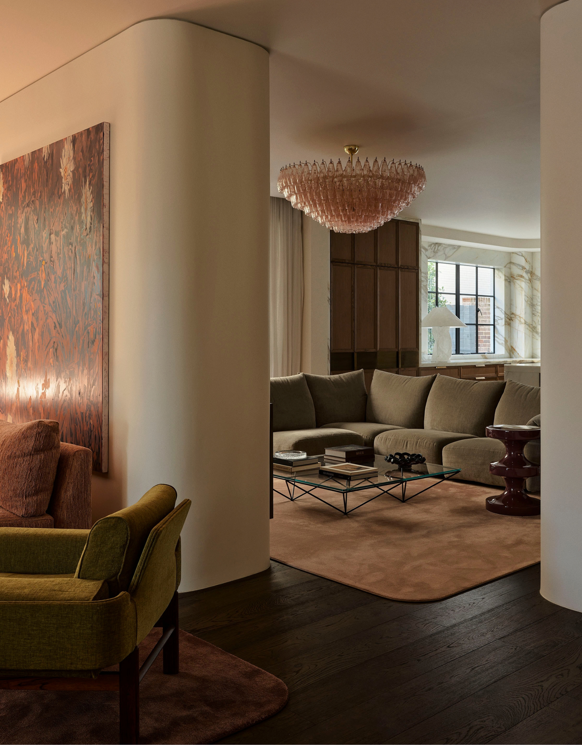 A modern living room with a large painting on the wall, curbed pillars, brown rugs, a green couch, a glass coffee table, and a pink chandelier