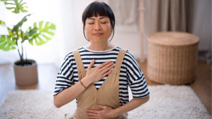 A woman kneels in a living room with her eyes closed, one hand on her chest and one hand on her belly. Behind her we see a wicker basket and leafy plant.