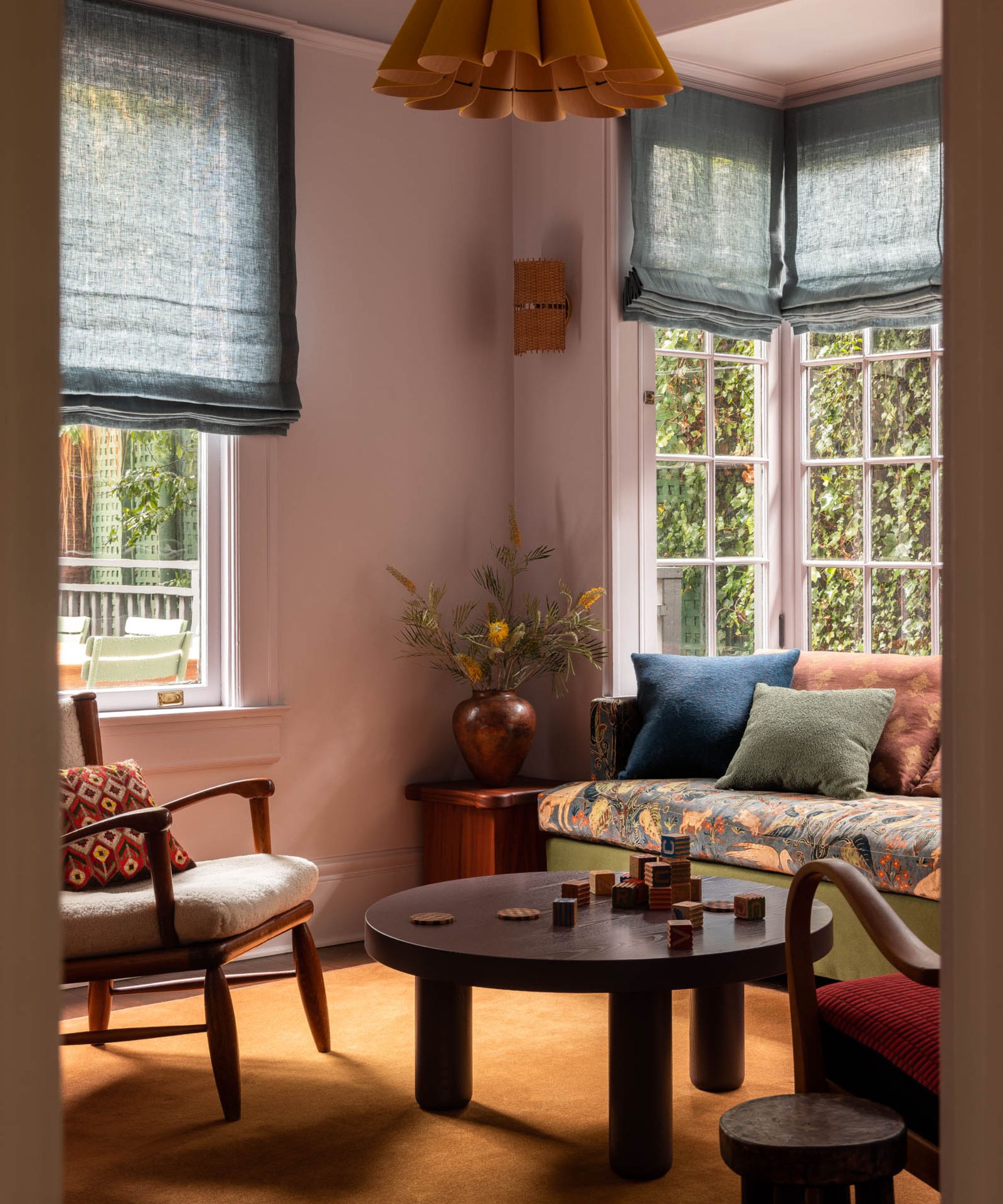 A living room with lilac walls, a bay window with blue Roman blinds, a sofa in the window with floral seat cushions and green, rust, and dark blue scatter cushions. An ochre rug, a modern coffee table, and an accent chair.