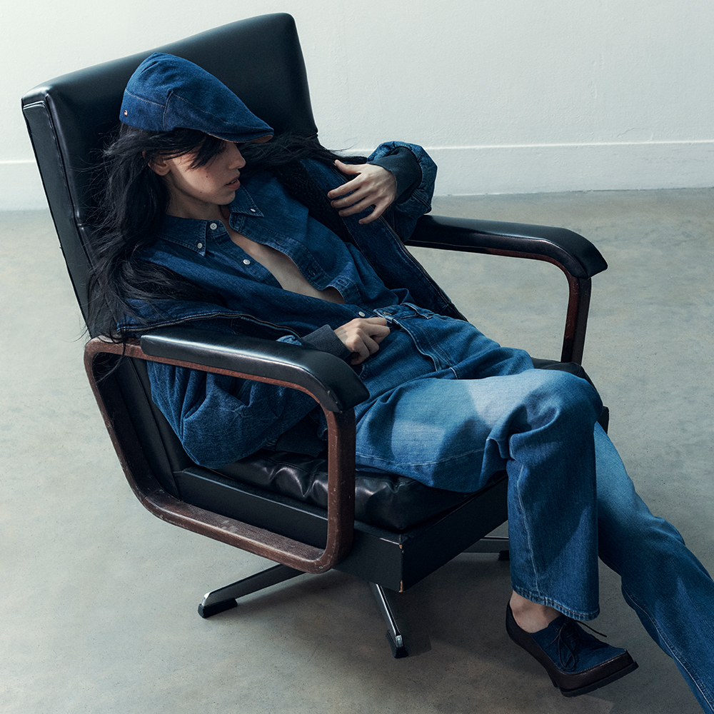 A photograph of a model reclining on a seat wearing blue jeans, a blue denim shirt and a blue denim hat