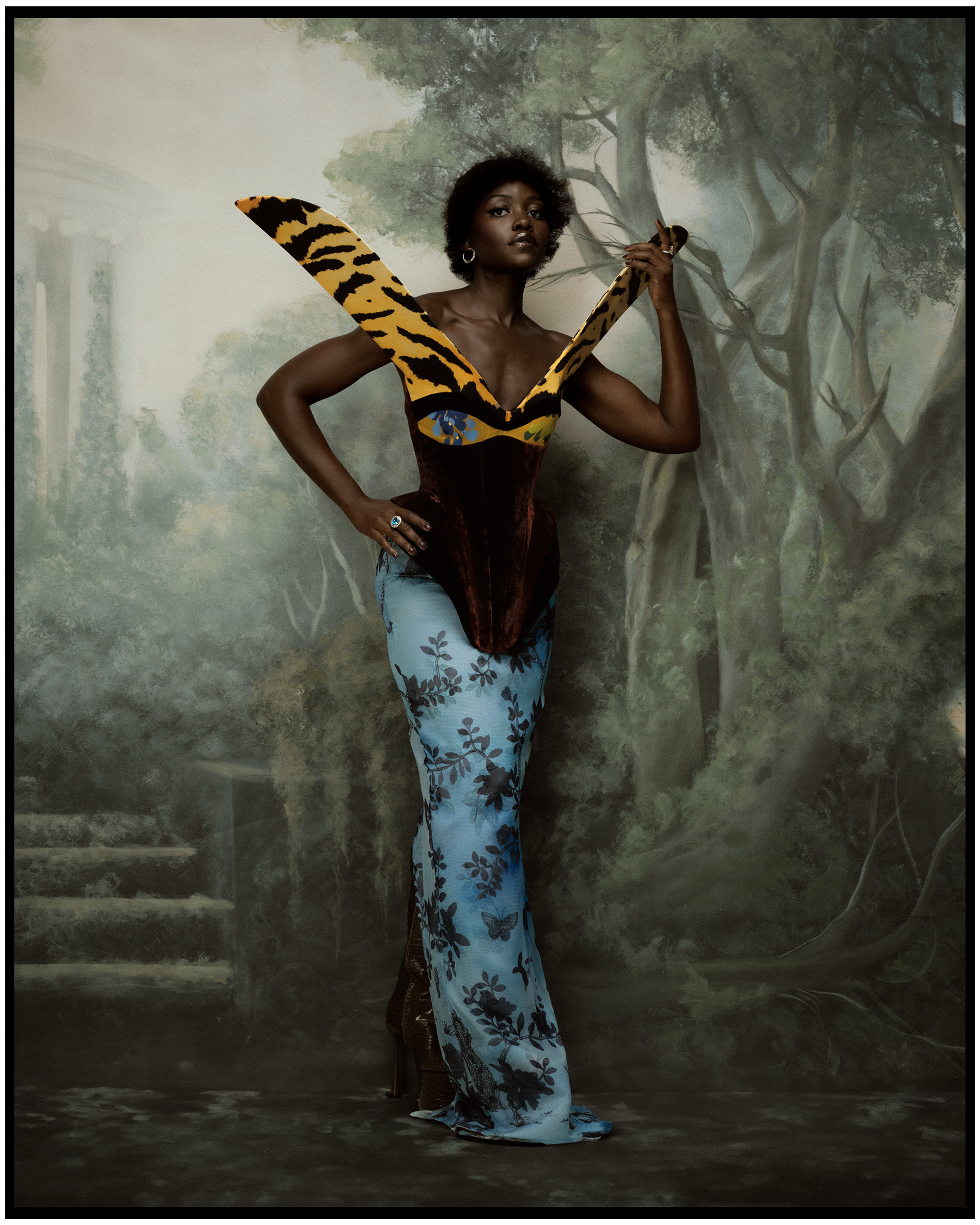 Lupita Nyong'o photographed by Kenny Germe&amp;#769; wearing a gown by Harris Reed. She is standing in front of a backdrop with a painted forest scene.