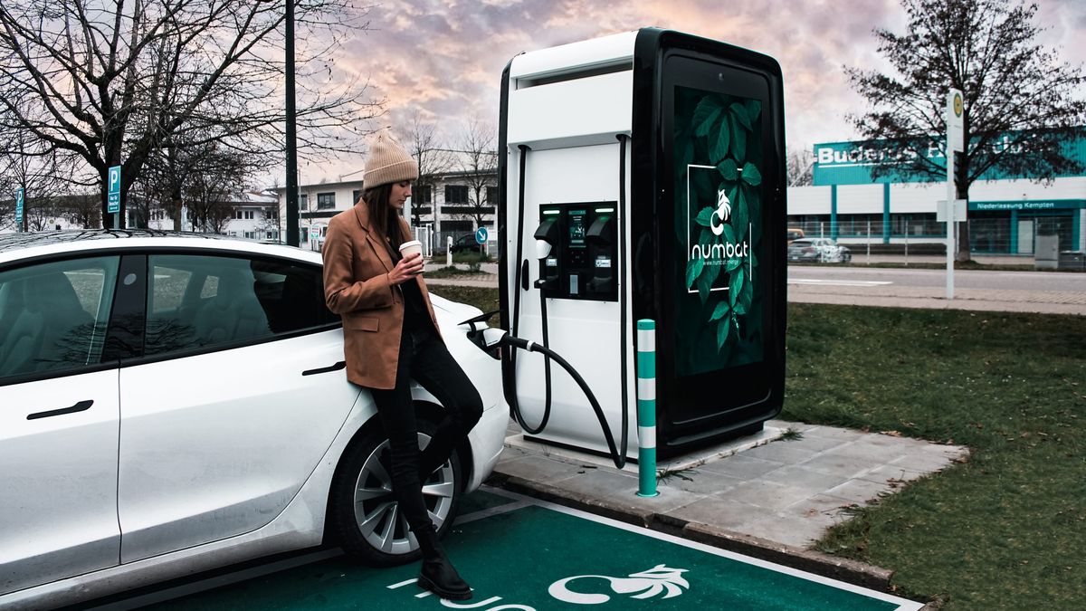 A woman leans on her car while charging her car at an EV station with DOOH advertising from Broadsign.