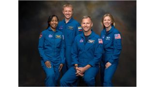 four smiling people in blue flight suits stand in front of a dark gray-brown background.