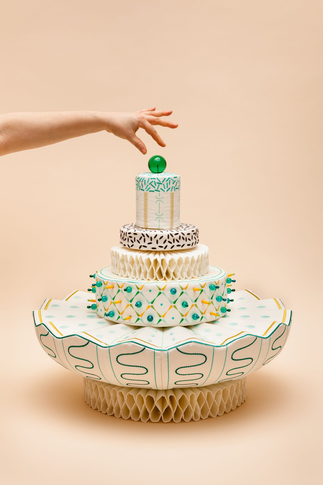 A cake-inspired sculptural installation in white, green, and yellow sits against a neutral beige background as someone pretends to place a green cherry on top of it.