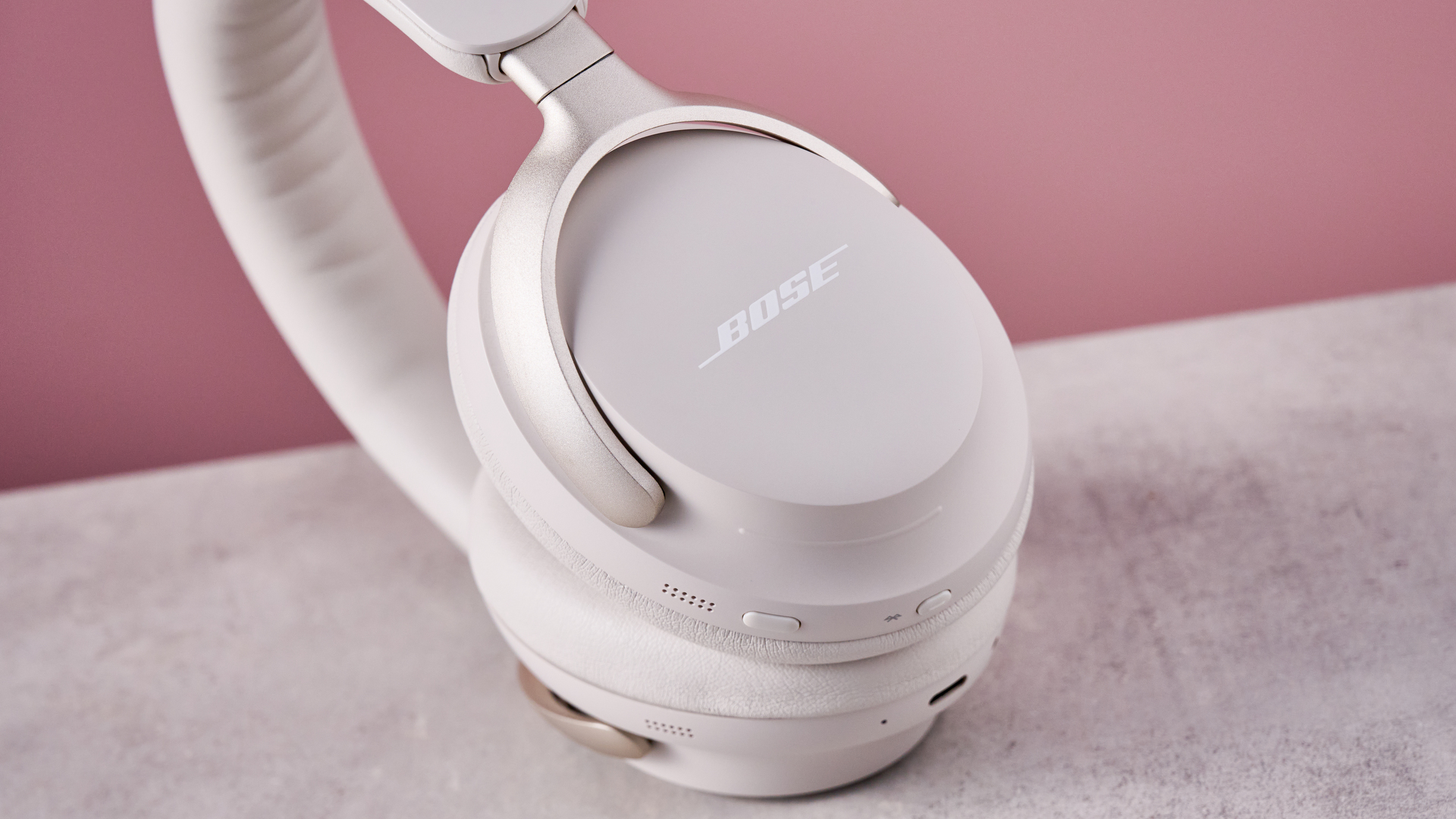 Bose QuietComfort Ultra Headphones on a gray surface with a pink background, showing the logo on the outside of the earcup