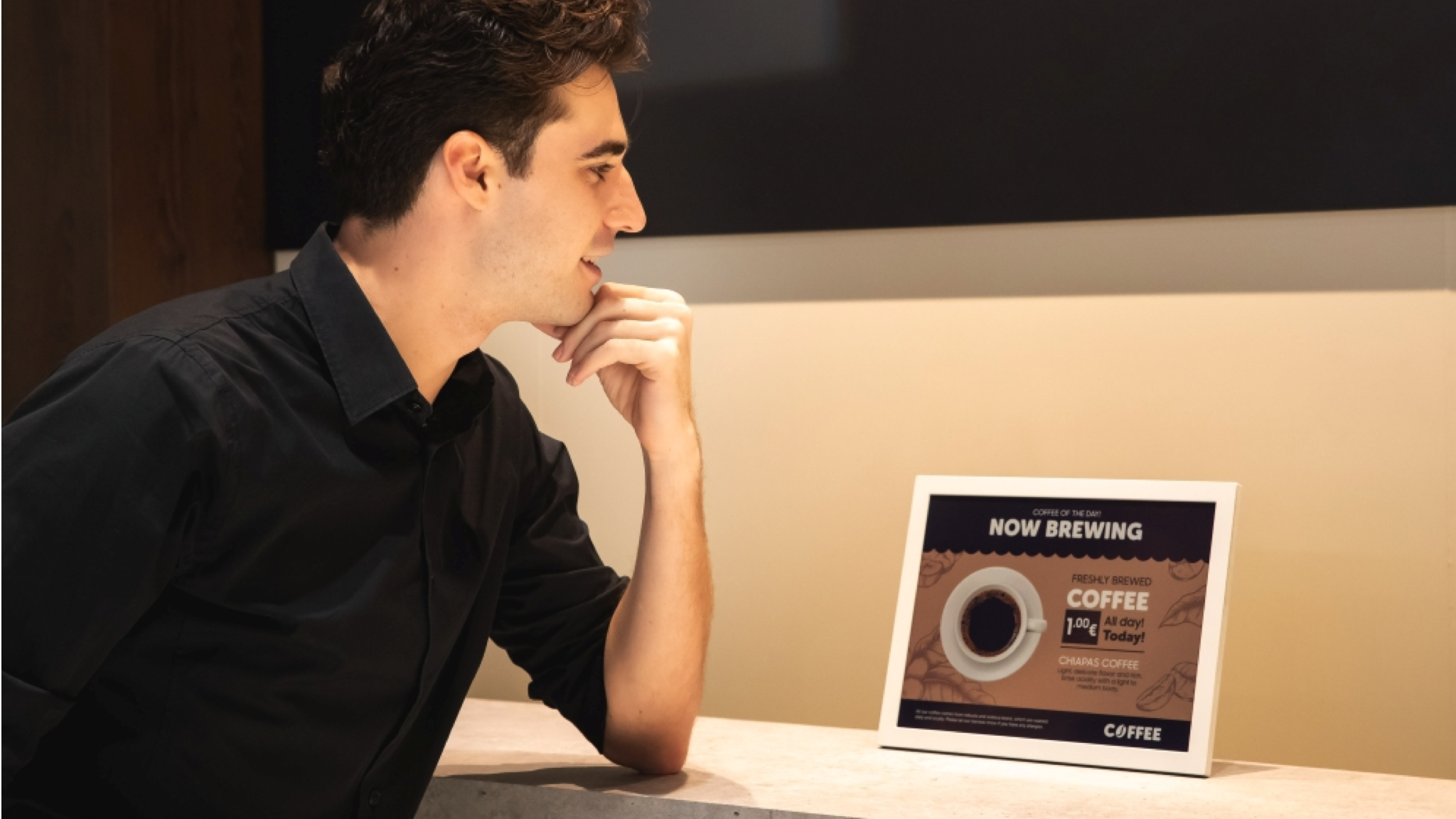 A man with dark hair and a black shirt sits looking approvingly at a Samsung Color E-Paper device showing a coffee advert