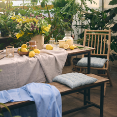 Wooden bench and chairs with neutral tablecloth, jugs of lemondade, bowls of lemons and a large bunch of flowers.