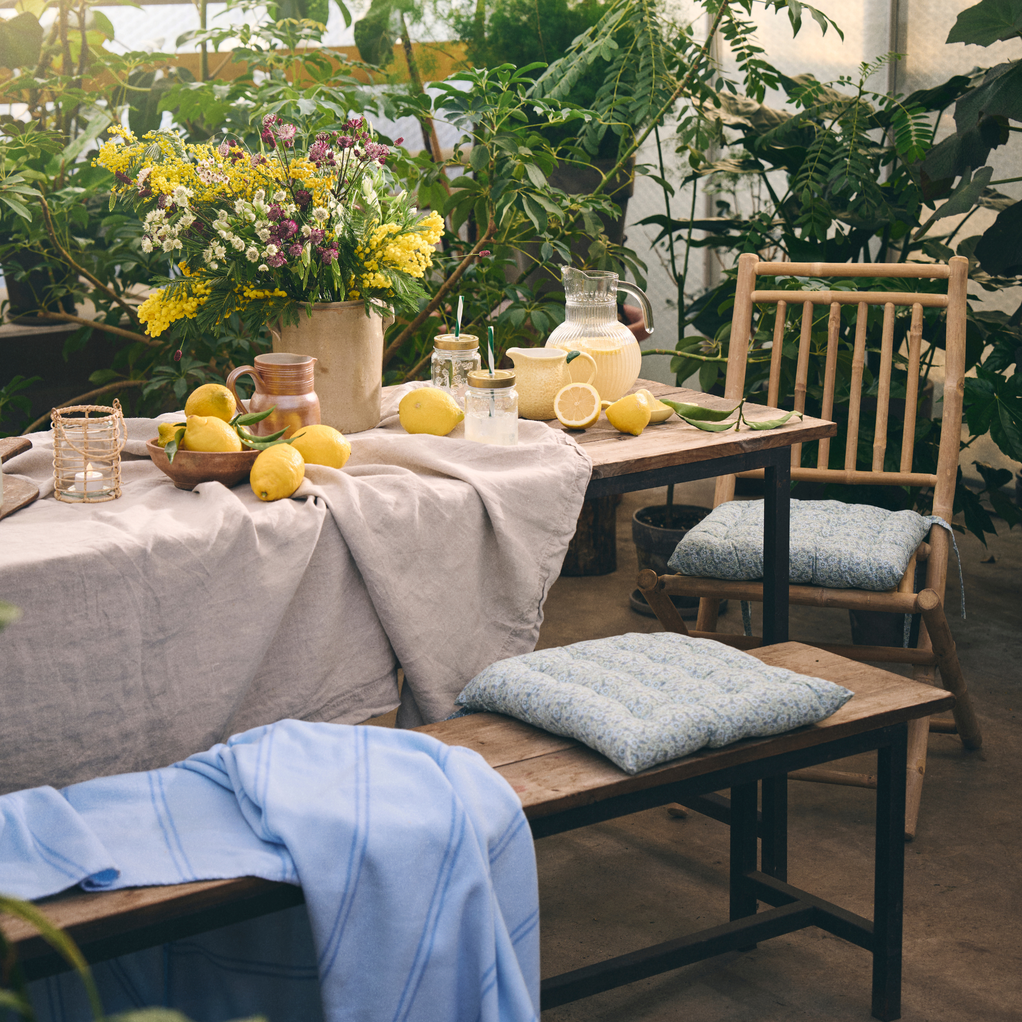 Wooden bench and chairs with neutral tablecloth, jugs of lemondade, bowls of lemons and a large bunch of flowers.