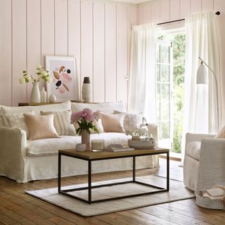 Soft pink living room with cream sofa with cushions in front of open window with white curtains