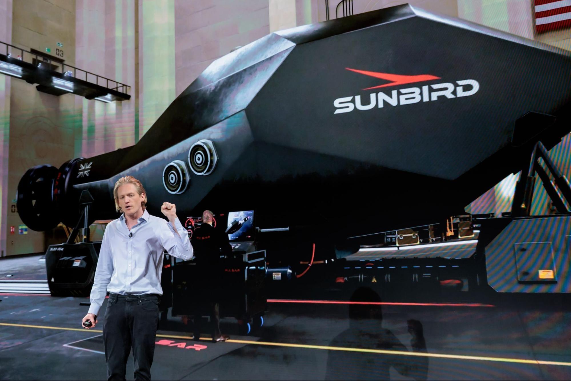 A man wearing a white shirt and black pants with blond hair stands next to a large black craft with the word "Sunbird" on it.