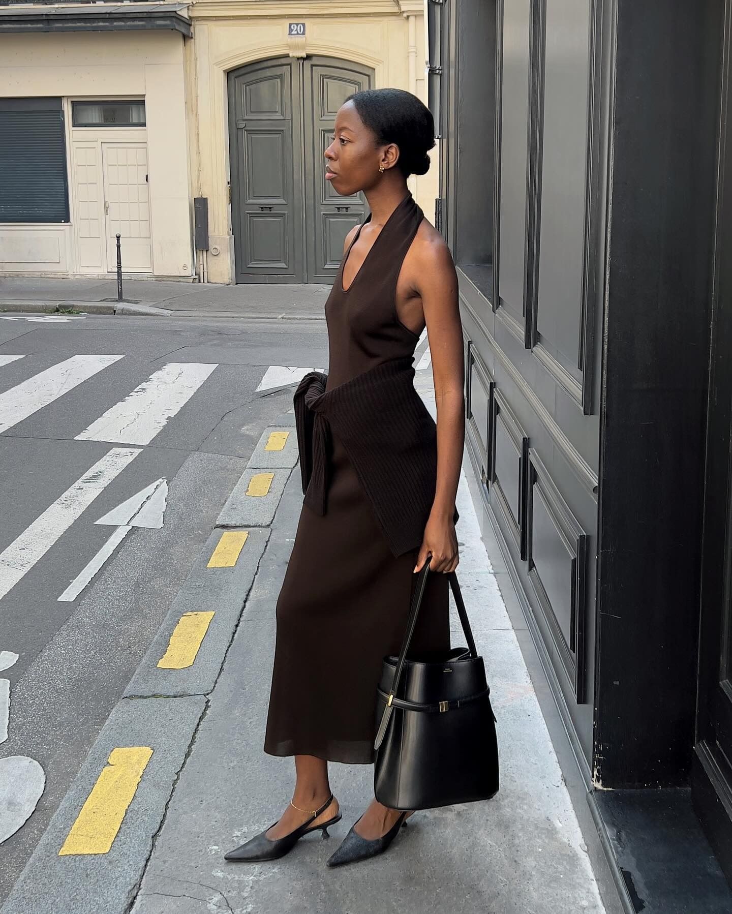 European style influencer Sylvie Mus poses on a sidewalk in Paris wearing a brown halter-neck dress with a sweater tied around her waist, a Toteme tote bag, and black slingback heels.