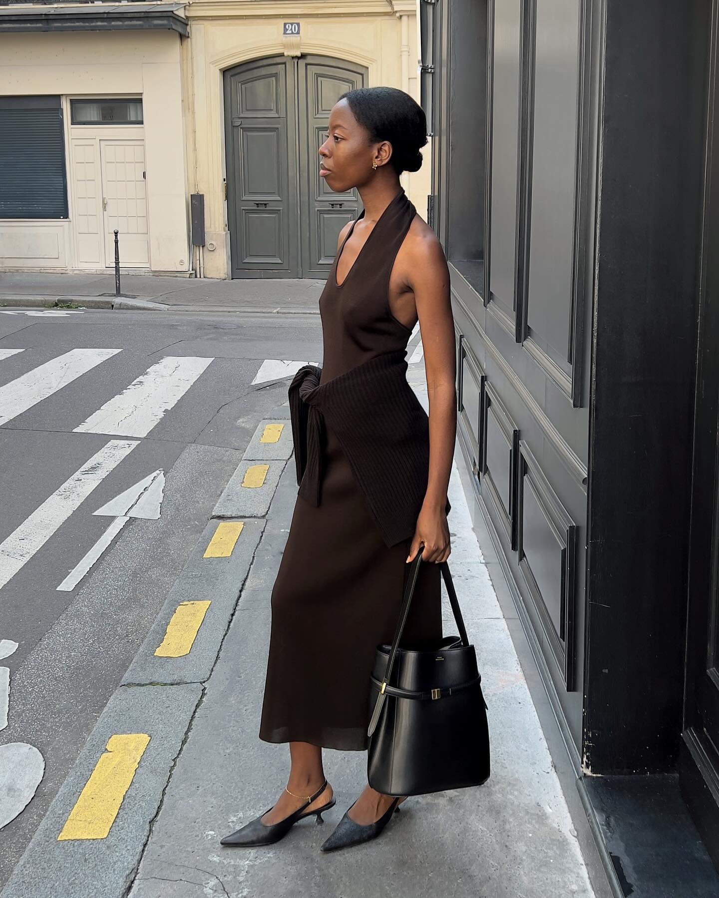 European style influencer Sylvie Mus poses on a sidewalk in Paris wearing a brown halter-neck dress with a sweater tied around her waist, a Toteme tote bag, and black slingback heels.