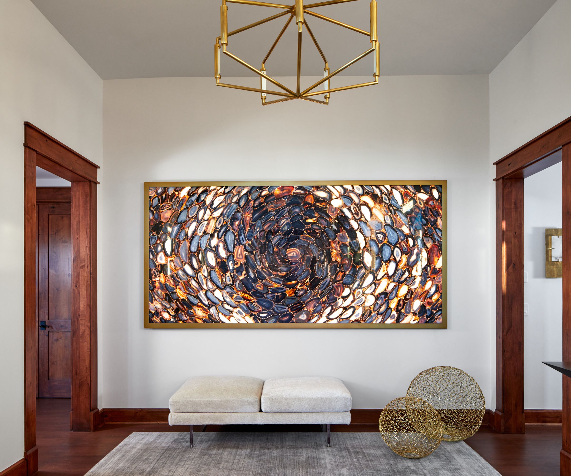 Modern entryway with large crystal artwork on back wall, bench, rug and two metal sculptures below, wooden flooring and door frames surround