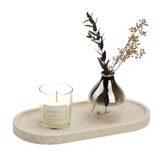 A light stone oval shaped tray with slightly lipped edges. A clear glass candle jar with white wax and an unburned wick on top next to a mirrored vase of faux dried stems and leaves.