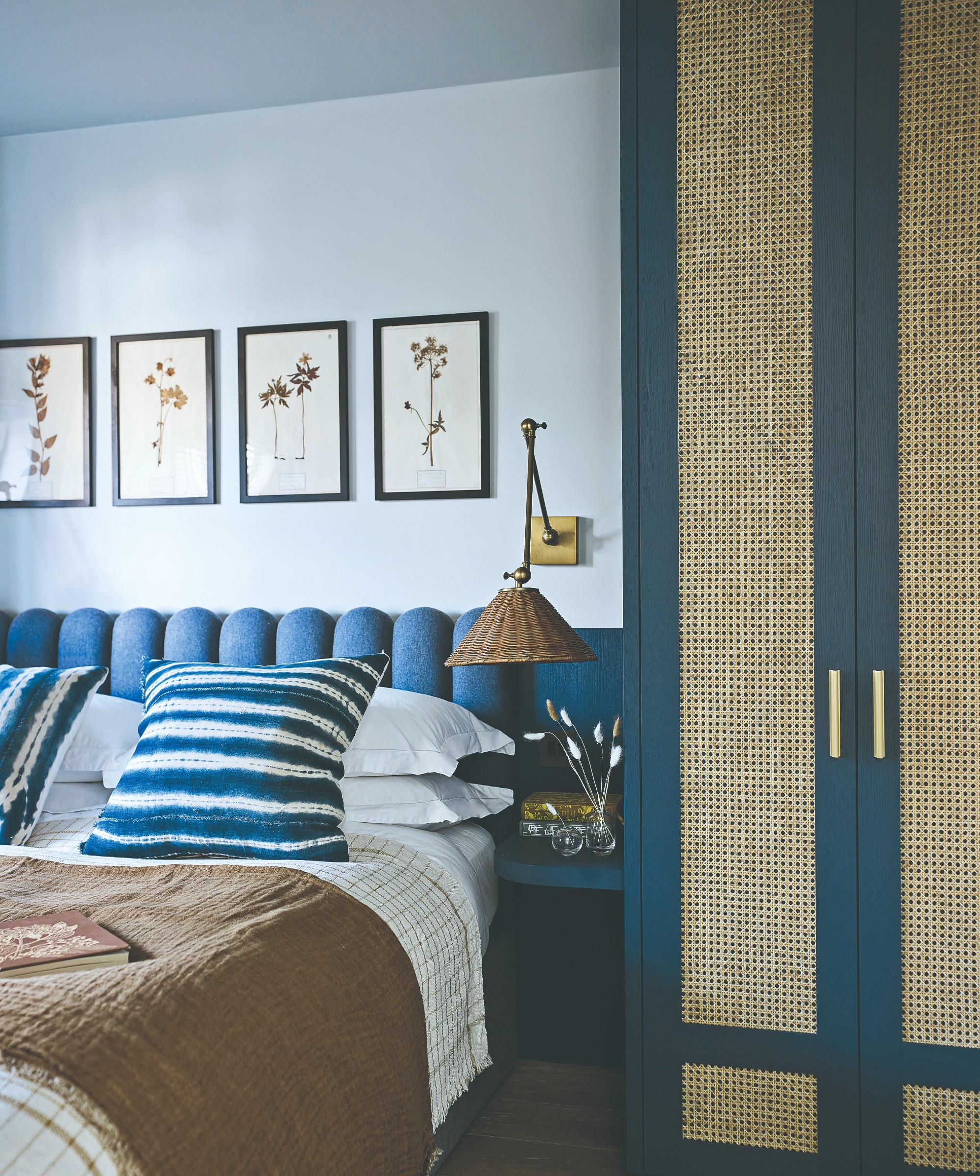 A blue bedroom with a scalloped headboard, a cane-insert wardrobe and a gallery wall of framed dried flowers