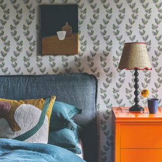 A bedroom with a botanical wallpaper and a bright orange bedside table next to a bed with a blue fabric headboard and blue bedding