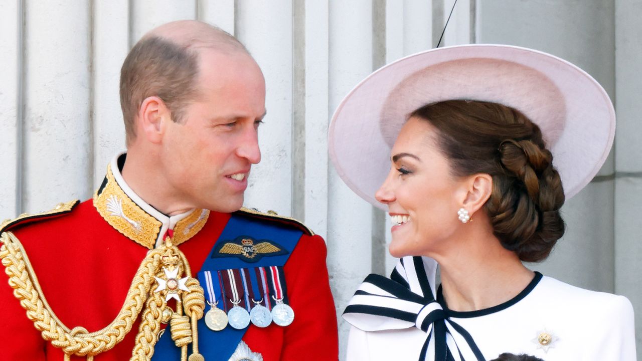 Prince William wearing a red military uniform with medals turning to look at Kate Middleton who is wearing a big white hat and white dress with a navy bow and smiling 