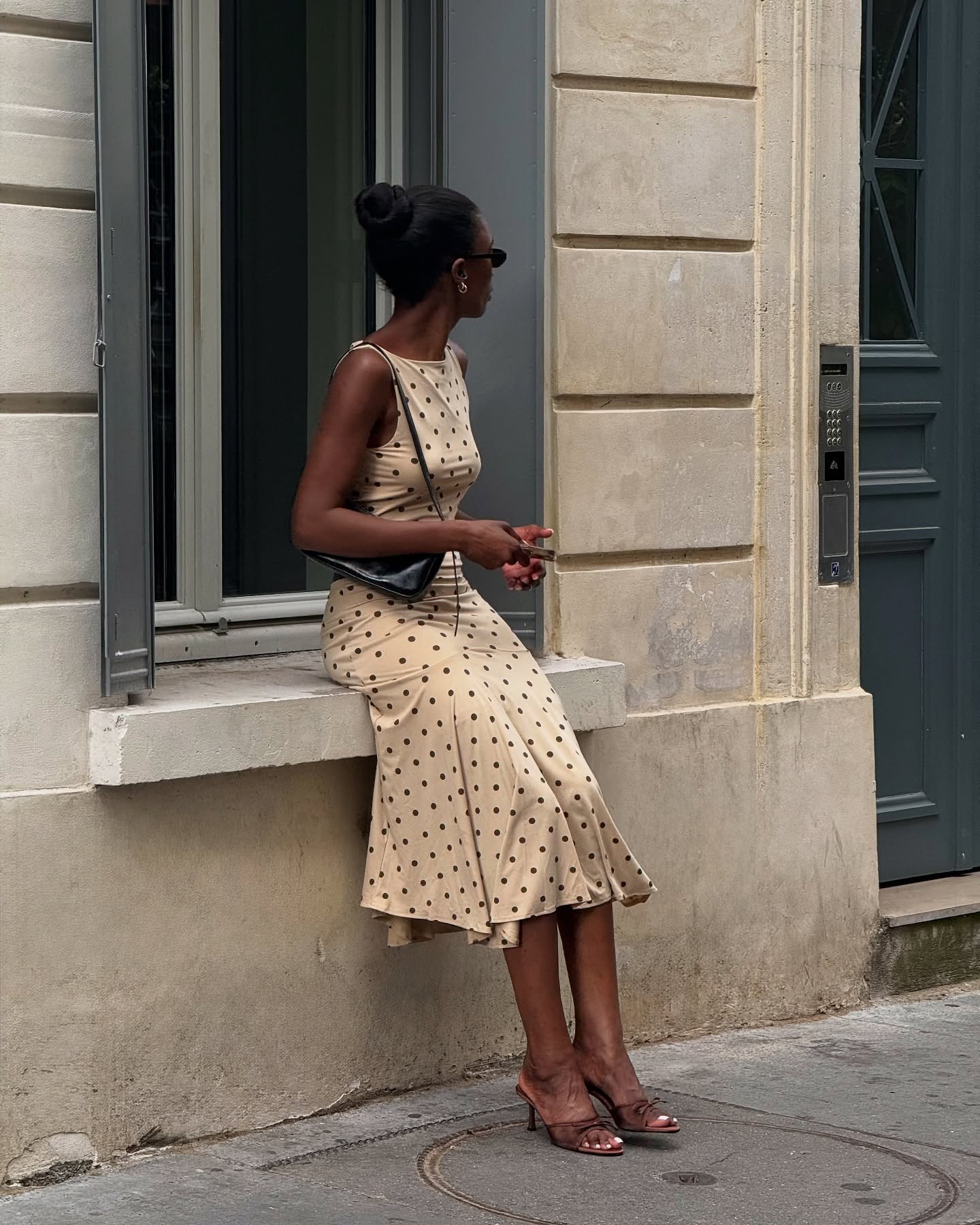 British style influencer Liv Madeline sitting on a window sill on a Paris sidewalk wearing black oval sunglasses, a black shoulder bag, a matching tan and black polka-dot skirt and top, and brown heeled mule sandals with a bow.