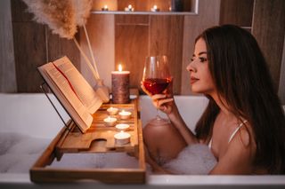 A young woman enjoying a bubble bath with the glass of red wine in hand, along with a bath tray that holds an open book and some candles.