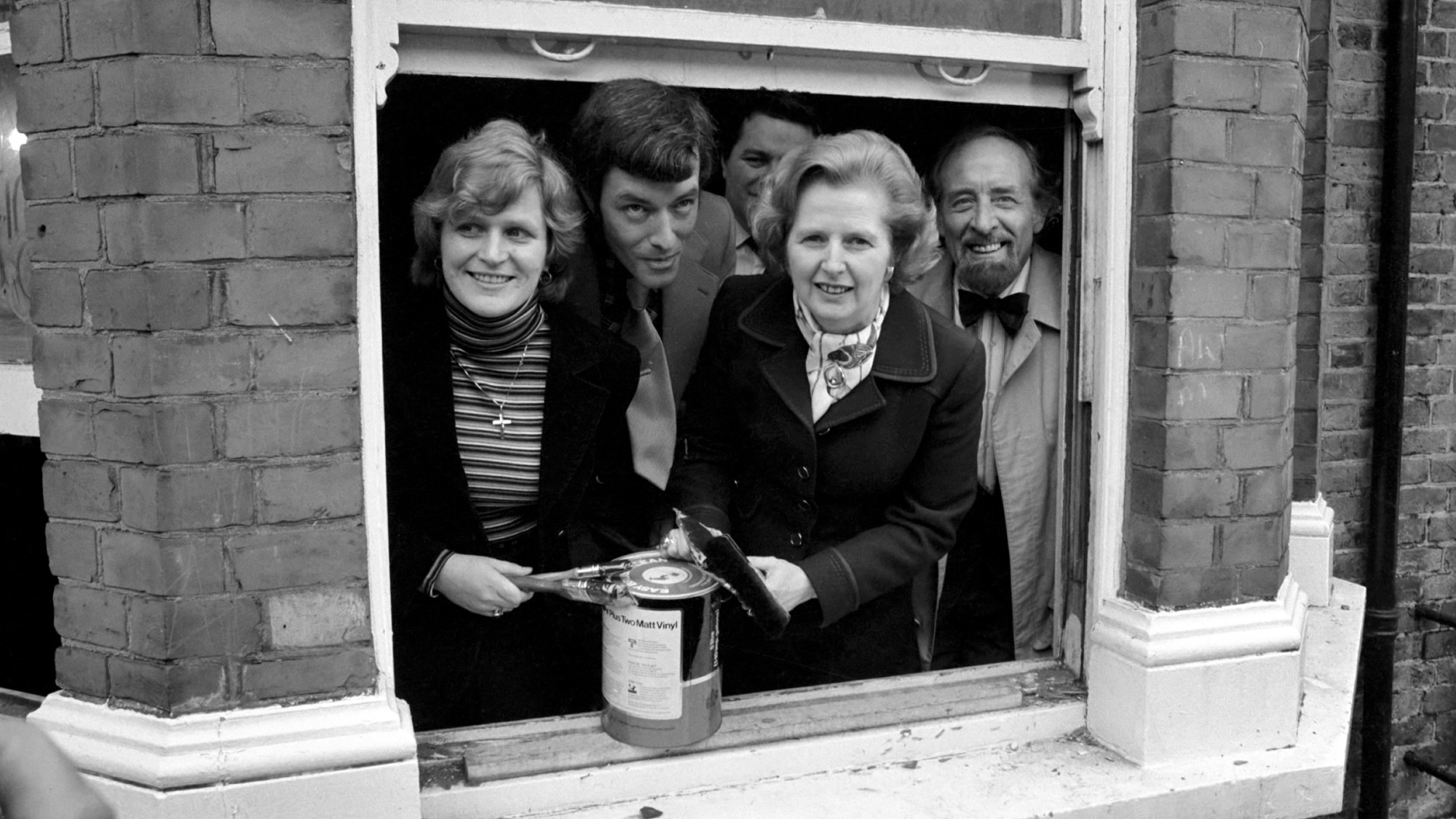 Margaret Thatcher opening a paint tin on the windowsill of an open window with a family around her