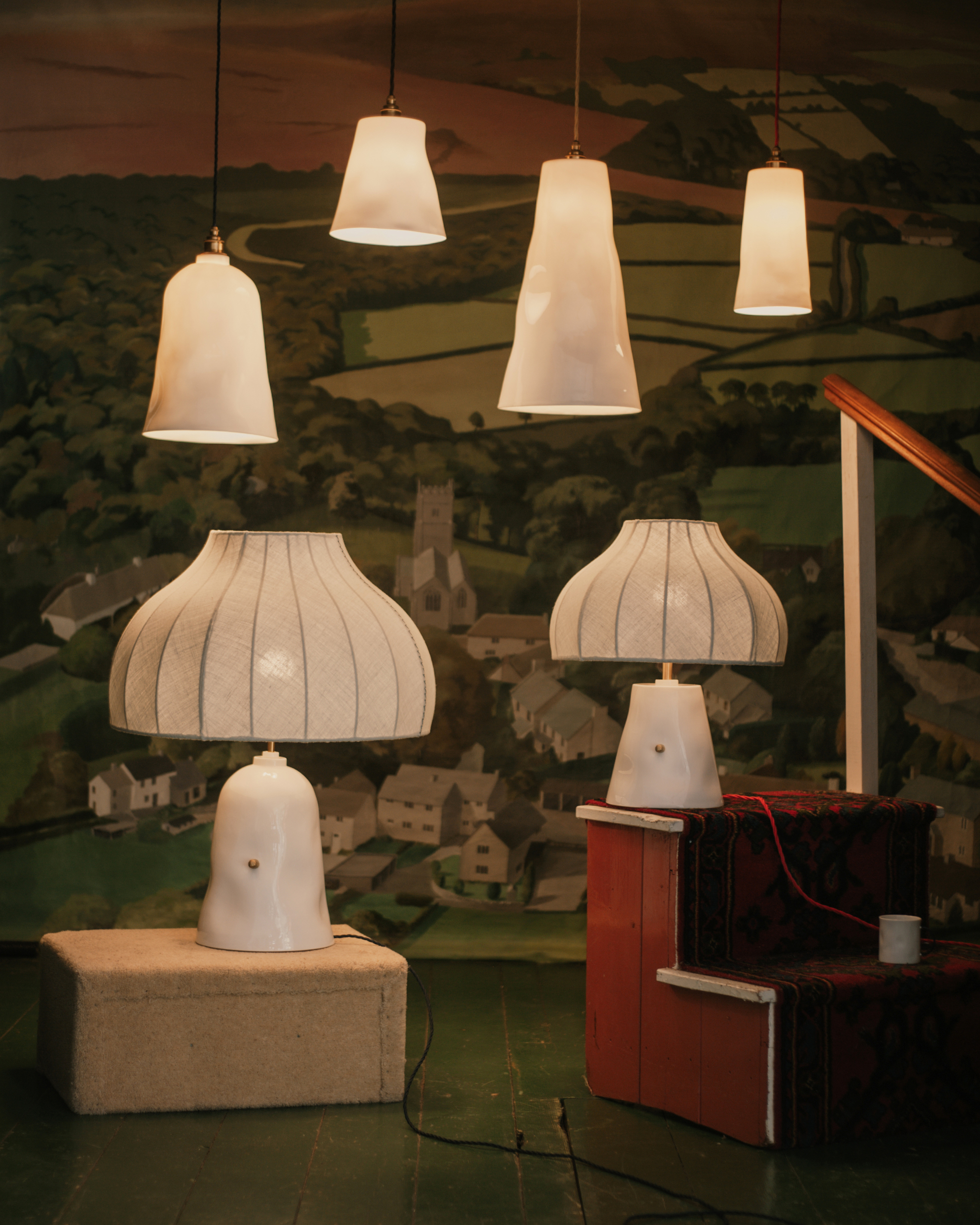 Image of two ceramic lamps and four ceramic pendant lights from lighting company Feldspar. They are styled on boxes in a room with mural wallpaper and green wooden floors.