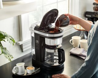 A female inserting ground coffee into drip machine demonstrating how to use a coffee maker in kitchen
