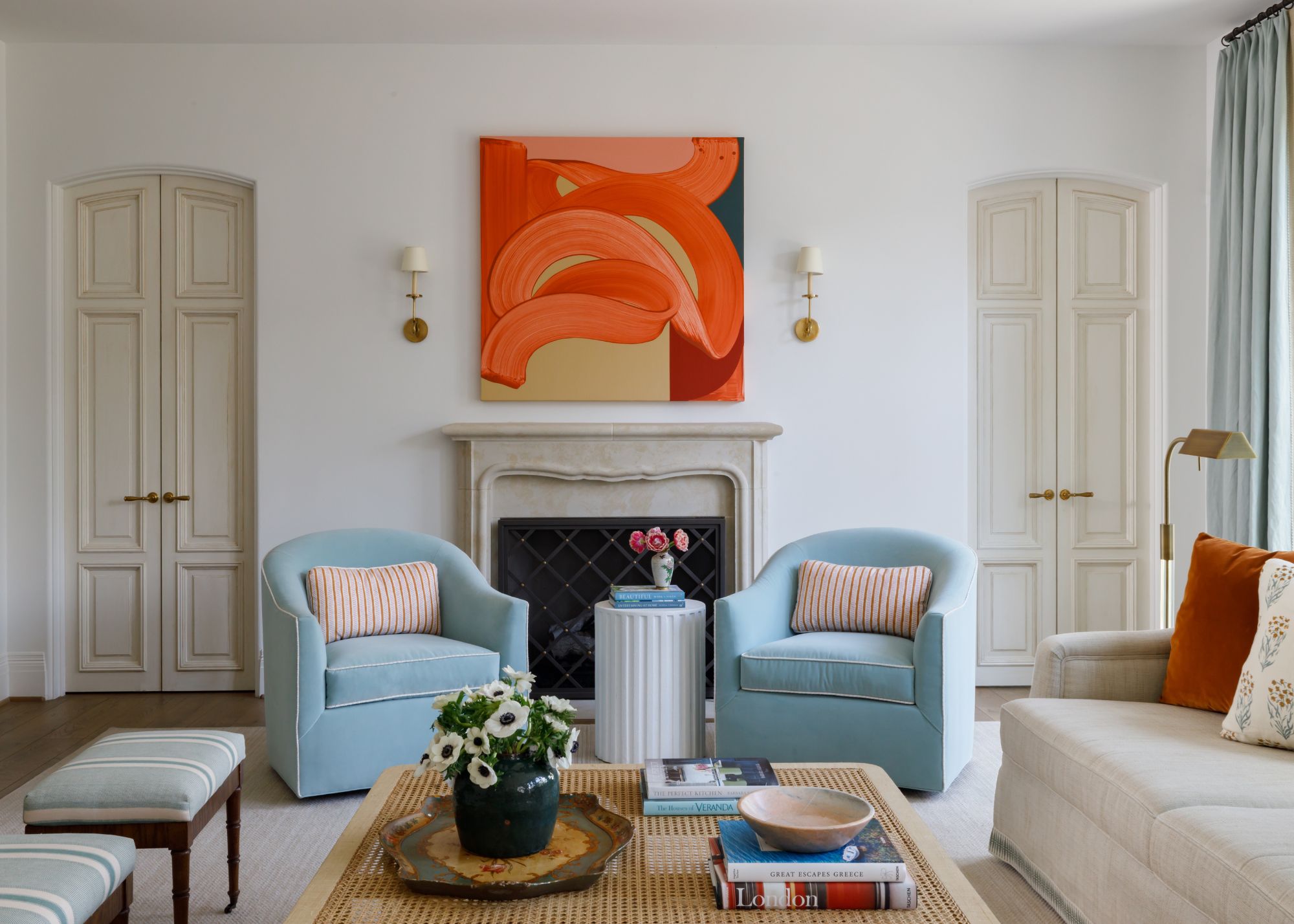 A large living room with white walls, two sets of doors leading into another room, a fireplace with a bright orange abstract artwork above it, and a cream sofa with two armchairs and a coffee table