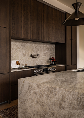 A dark, modern kitchen with a leathered grey marble waterfall edge island and a matching backsplash the cabinetry is in a dark wood