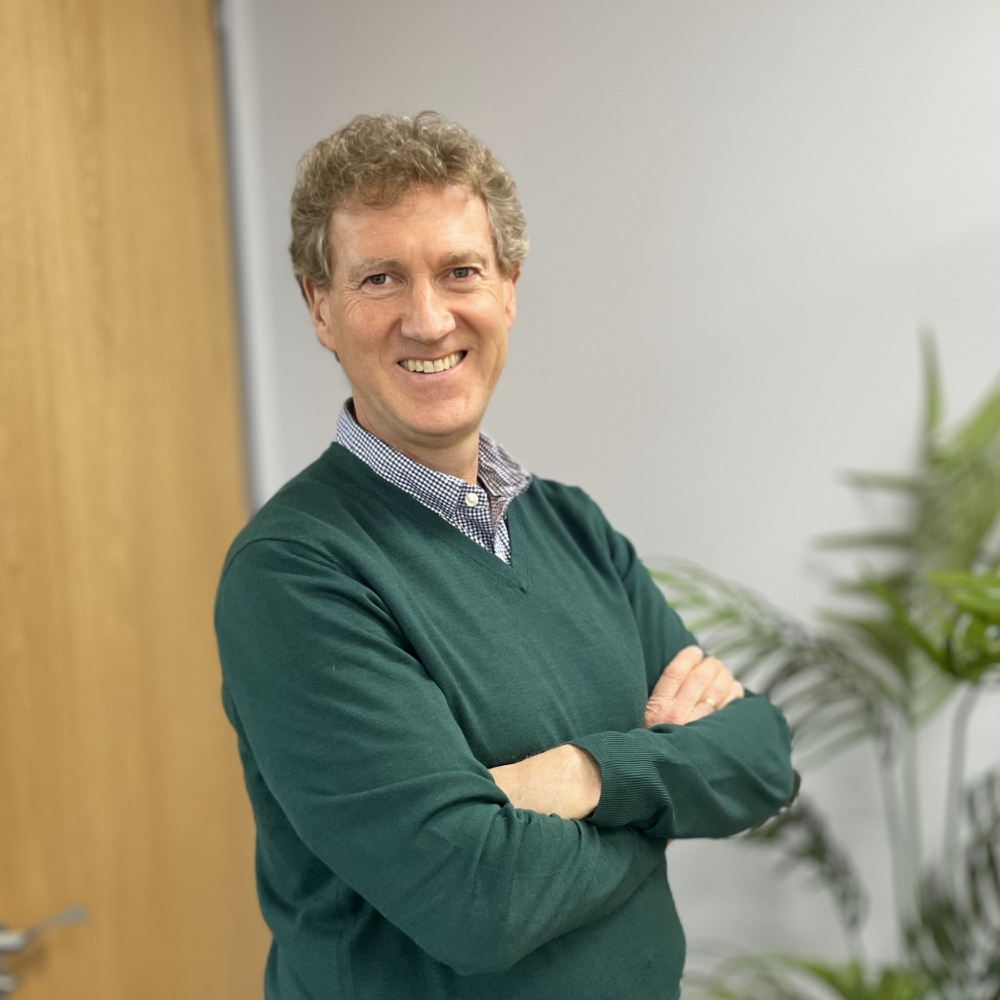 male with short dark blond hair stood in office wearing green v neck jumper and check shirt