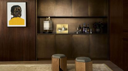 A brown bar room with bronze cabinets, a hanging painting, and a pair of pouffes.