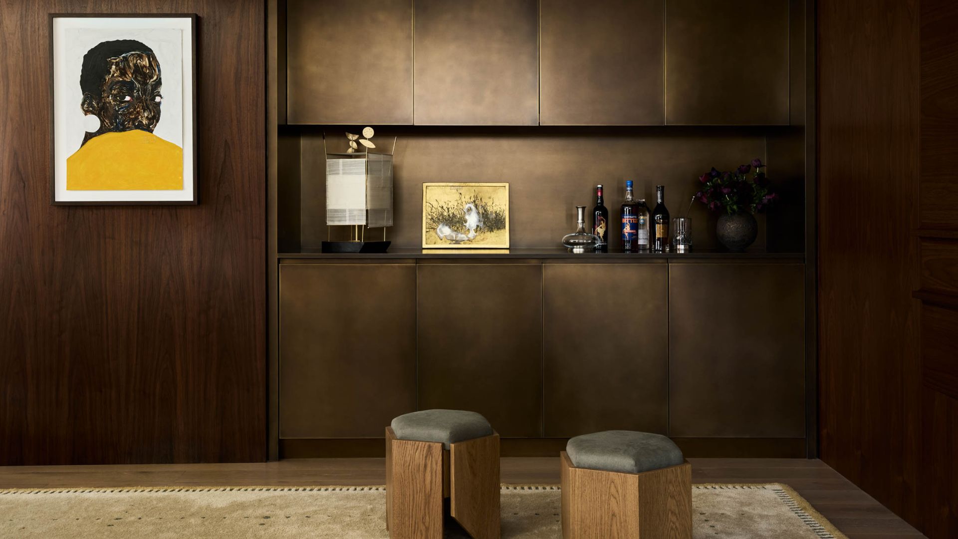 A brown bar room with bronze cabinets, a hanging painting, and a pair of pouffes.