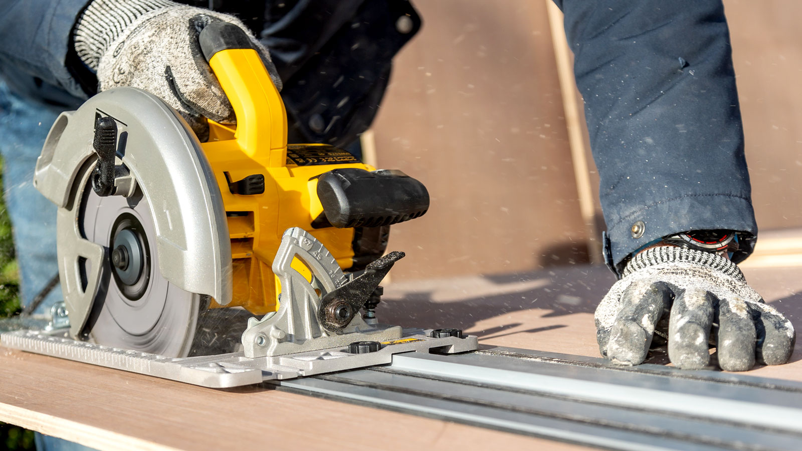 Yellow circular saw held by person in gloves using a guide rail to cut a sheet of plywood
