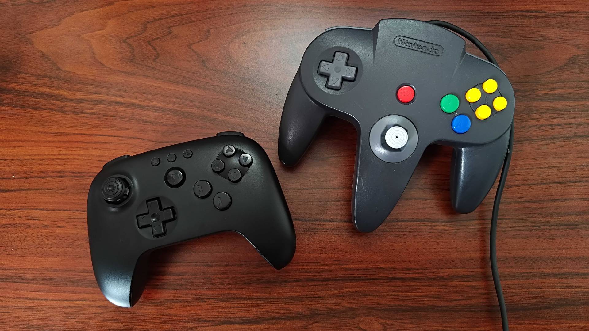 8Bitdo N64 Bluetooth controller next to original black gamepad on woodgrain surface.