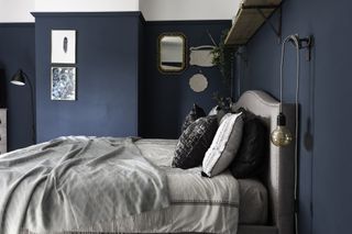 Deep blue bedroom with white ceiling paint that carries down wall, grey upholstered bed, monochrome bed covers, exposed bulbs as bedside lights