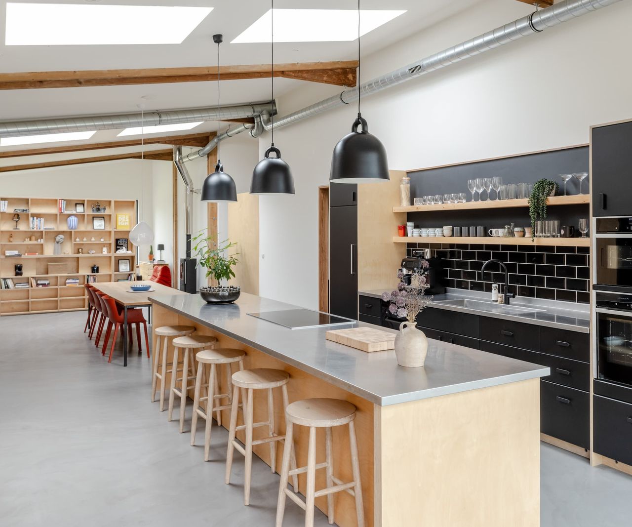 An open plan kitchen with a long kitchen island with an induction hub and overhead rooflights and a dining area with black cupboards