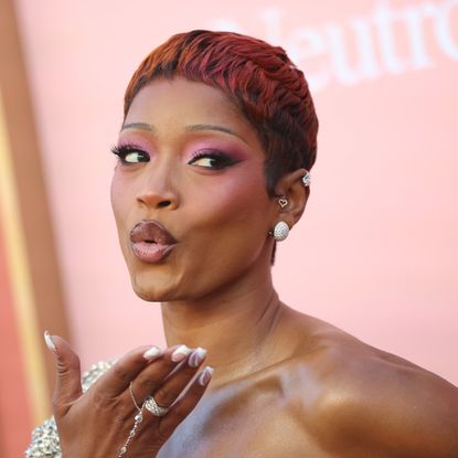 a black woman with short red hair wears pink eyeshadow and poses in front of a peach background