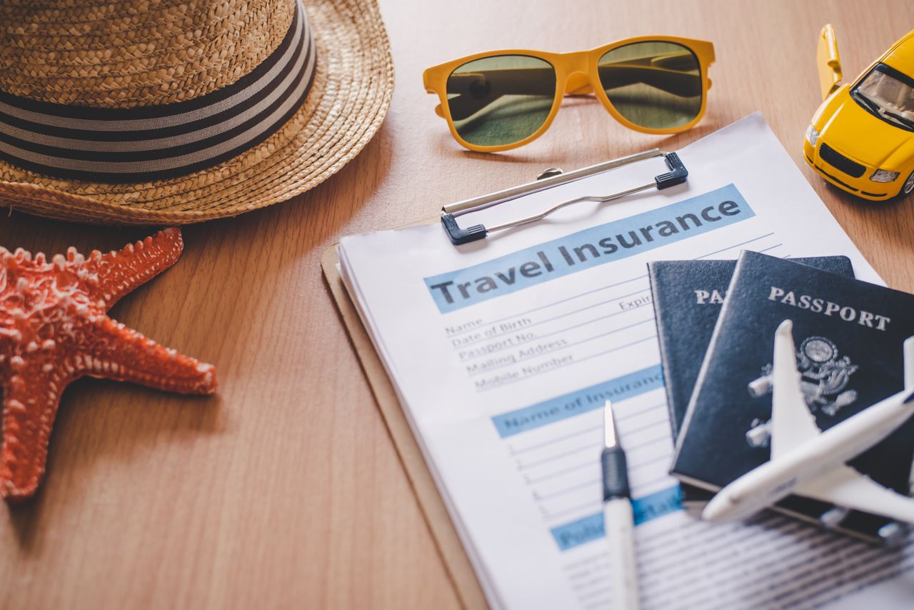 Travel insurance document on clipboard beside passports, pen, sunglasses and hat among other items.