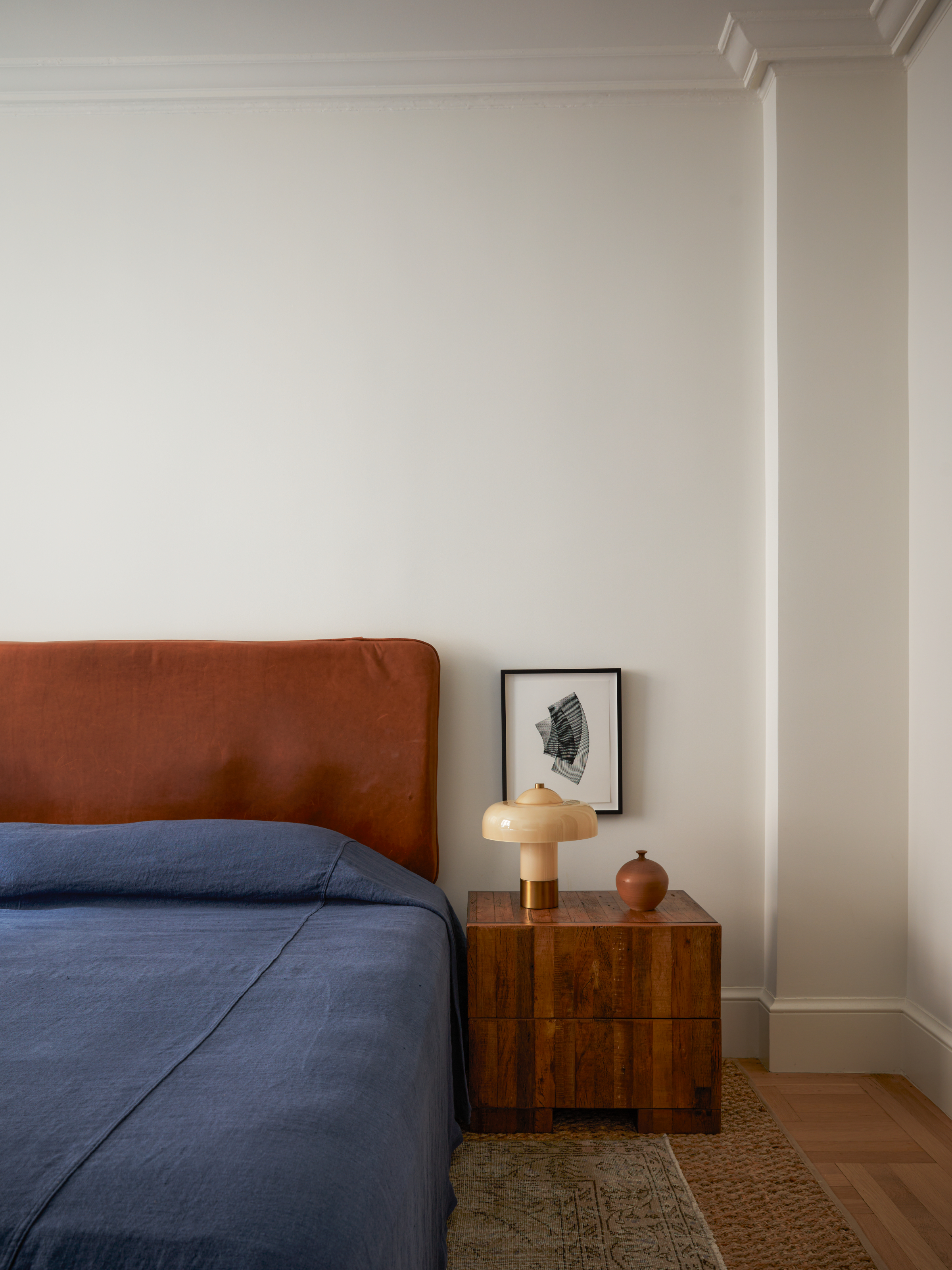 Bedroom with tobacco-colored upholstered headboard, cherry wood side table, navy bed linen and cream lamp