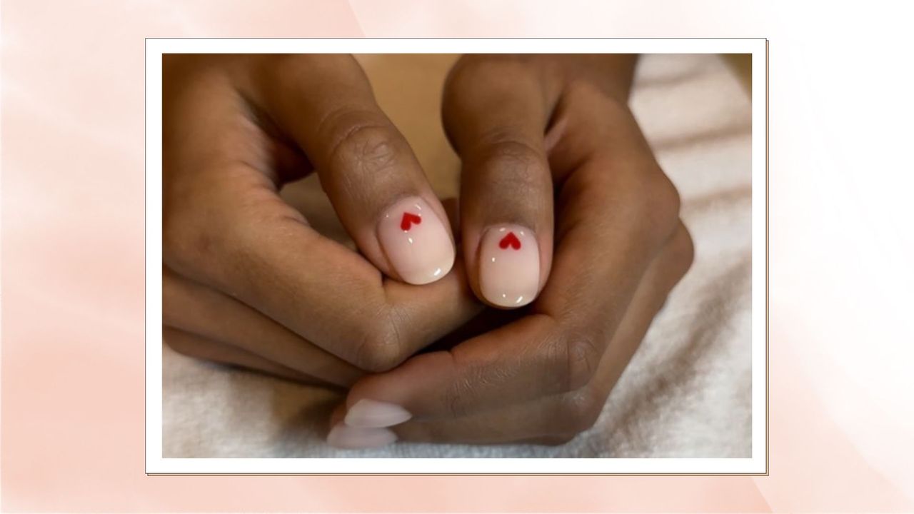 An image of a womans hands folded on a towel with cream Biab nails with a little red heart on them