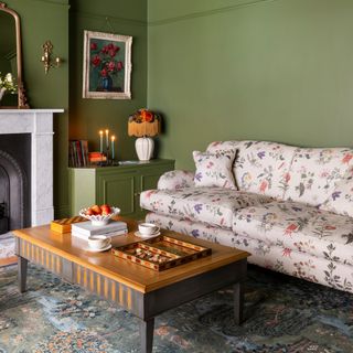 A green-painted living room with a floral-print sofa and a rectangular wooden coffee table with two coffee table books, a fruit bowl and board games