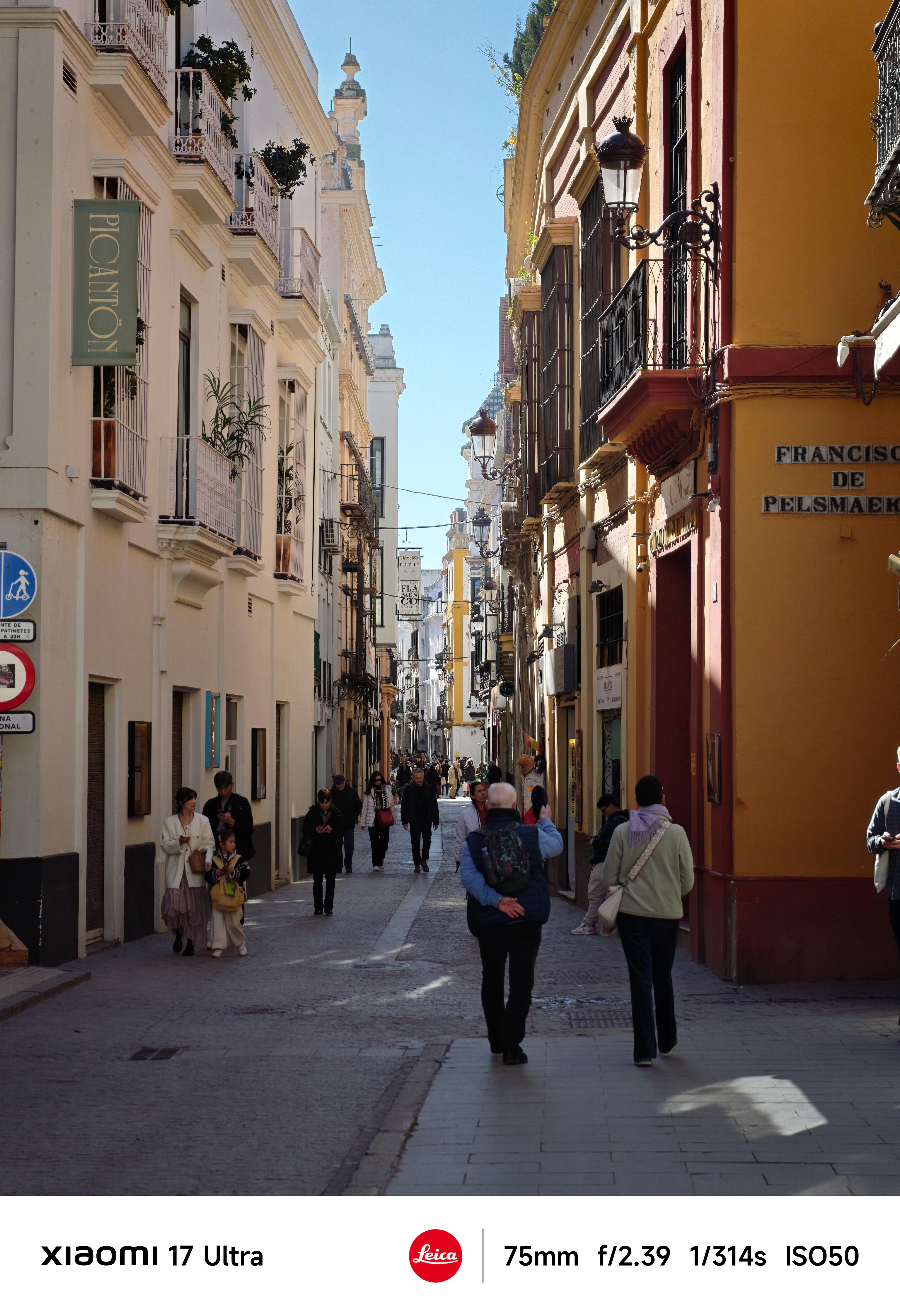 Narrow European street lined with pastel buildings and wrought-iron balconies, pedestrians walking in bright midday sun.
