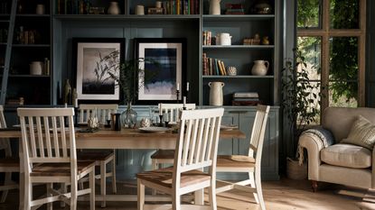 A dining toom with large dark green built in shelves on the back wall beside a window. An upholstered armchair beside it. A white and wood rectangular dining table in front of it with matching chairs surrounding it. 