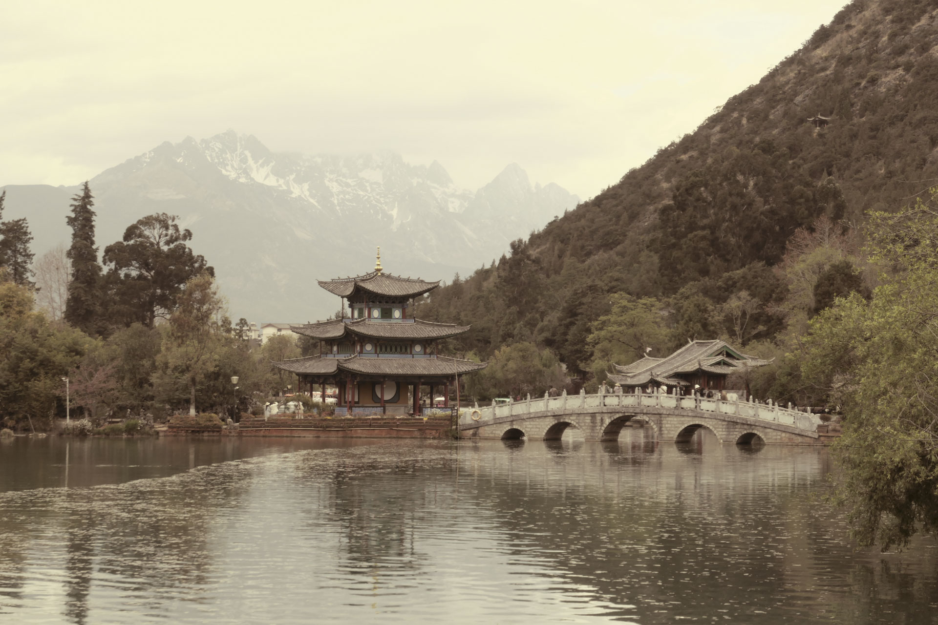 A Chinese temple by the side of a lake with an arched bridge leading up to it and both are reflected in the still water, in front of a snow covered mountain. A creative filter is applied to the image