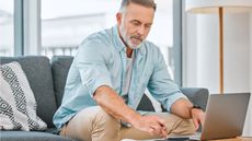 An older man uses a calculator and a laptop while sitting on his sofa.