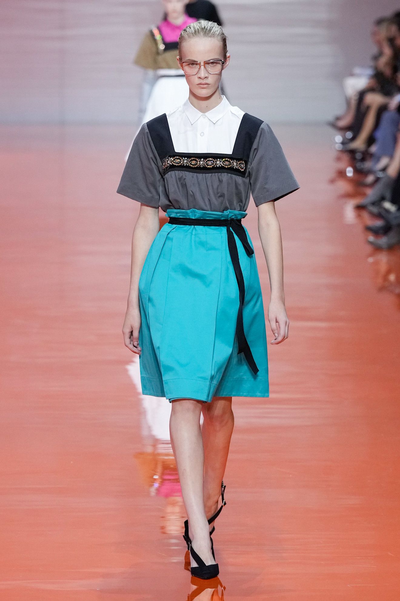 A model walking in the Prada SS26 show wearing a white button-down shirt under a square-neck bejewelled top with a turquoise satin skirt.