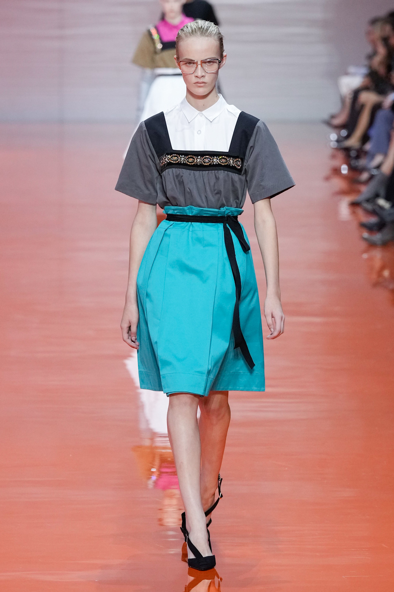 A model walking in the Prada SS26 show wearing a white button-down shirt under a square-neck bejewelled top with a turquoise satin skirt.