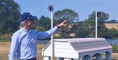 Derek di Grazia, wearing a light blue shirt and a navy cap, stands in front of a pink and white cross-country fence in Burghley Park. His arm is raised as he talks about the combination.