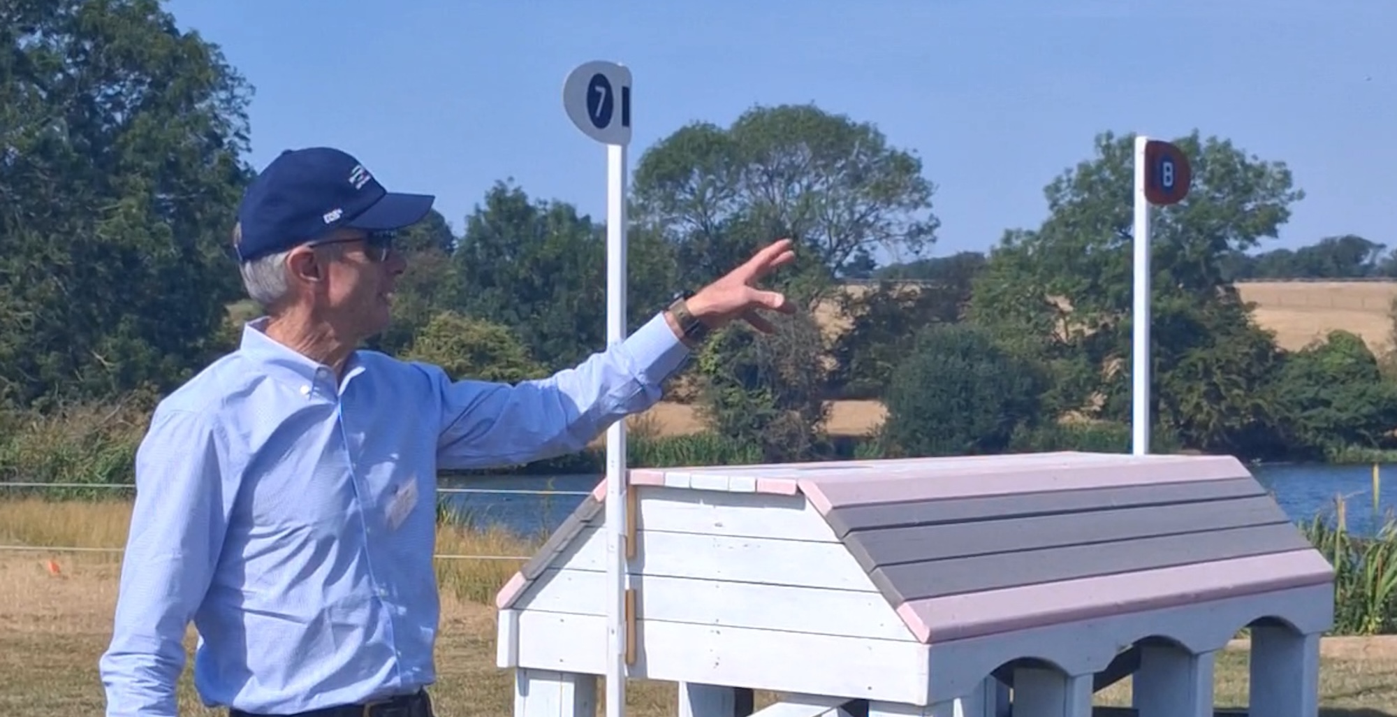 Derek di Grazia, wearing a light blue shirt and a navy cap, stands in front of a pink and white cross-country fence in Burghley Park. His arm is raised as he talks about the combination.