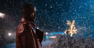 The Doctor (Ncuti Gatwa) stands in profile wearing a long leather coat. Snow is falling all around him and there are Christmas lights in a tree in the distance.